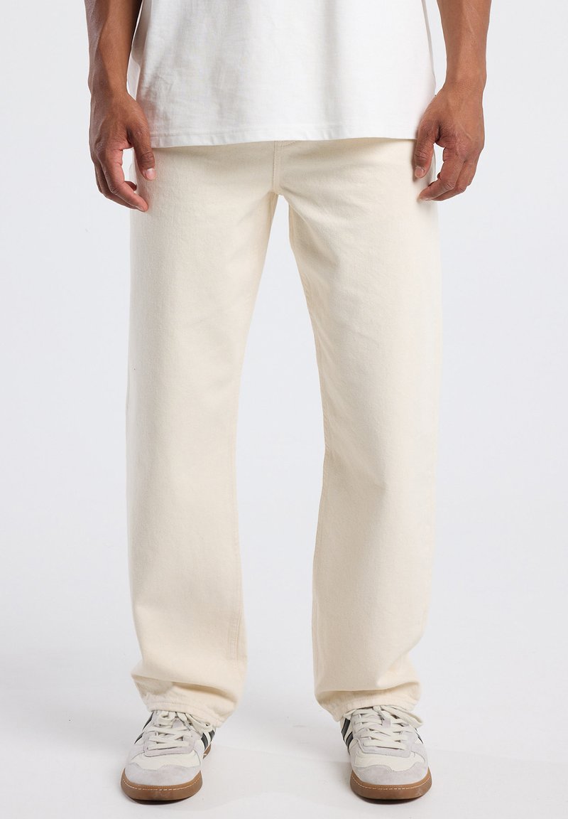 Hombre vestido con pantalones rectos color blanco roto, zapatillas blancas con rayas negras y una camiseta blanca, de pie frente a un fondo liso.