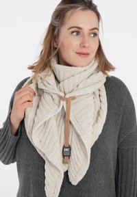 Beige gebreide sjaal met een driehoekig ontwerp, voorzien van een gestructureerd patroon en een bruine stoffen label, vastgebonden met een bijpassende band.