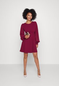 Bordeaux kjole med lange puffermarmer, figursydd midje og vide skjørt; modell som holder en metallisk clutch; nøytrale hæler; ensfarget bakgrunn.