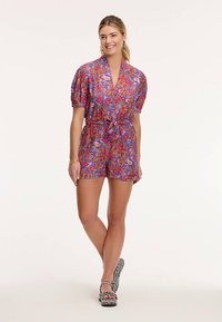 Krāsains ziedu raksta kombinezons ar īsām pūkainām piedurknēm, jostu viduklī un šortiem; izgatavots no viegla auduma; valkāts ar rakstainām sandales.