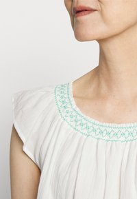 Blusa branca com um decote redondo adornado com bordado turquesa, feita de um tecido leve e suave, e com mangas em forma de campainha.