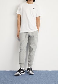 Person som bär en vit Nike-t-shirt, grå Nike-sweatpants och svartspräckliga Nike-sneakers som står på en blå matta mot en vit vägg.