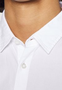 Biała bawełniana koszula z kołnierzykiem, zapinana na standardowe guziki, z widocznym przeszyciem i gładką teksturą wokół szyi.