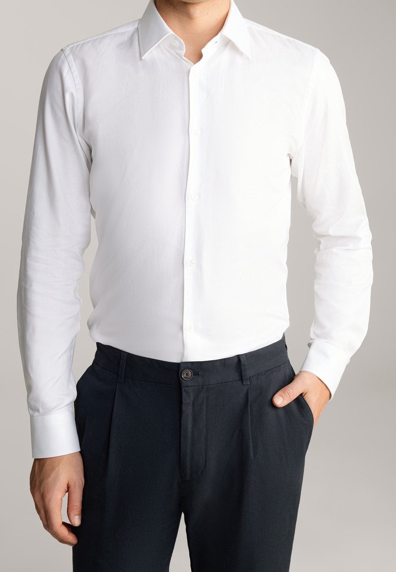 Homme portant une chemise blanche ajustée à manches longues boutonnée, rentrée dans un pantalon bleu marine foncé, une main dans la poche, sur un fond neutre.
