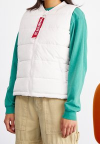 Weißer Steppweste mit hohem Kragen, Reißverschluss und roten Tagdetails, getragen über einem türkisfarbenen Langarmshirt und khakifarbenen Cargo-Hosen.