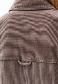 Bruine corduroy jas met verticale strepen, voorzien van een kraag en een lus als hangdetail aan de achterkant. Zachte textuur en een relaxte pasvorm.