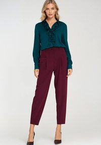 Niebieskozielona bluzka z guzikami z przodu z falbaniastym detalem, zestawiona z wysokimi bordowymi spodniami. Modelka nosi czarne, spiczaste czółenka.
