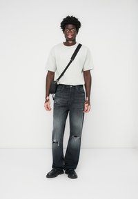 Grå t-skjorte med kort erme, slitte mørke jeans med hull, svart skulderveske med stropp og svarte sko. Modellen har krøllete svart hår.
