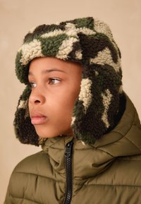 Camo-patterned, fleece-lined hat with ear flaps in green, brown, and cream. Worn with a zippered olive green puffer jacket.