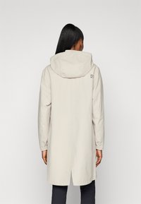 Imperméable beige clair avec capuche, doté de poignets élastiques, d'une coupe droite et d'un petit logo au dos. Matière lisse et résistante à l'eau.