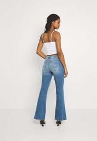 Jeans de ganga azul com cintura alta e boca de sino. Apresenta dois bolsos traseiros e costura discreta. Combinado com uma blusa branca justa tipo camisola.