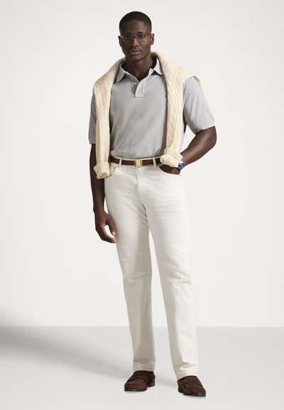 Γκρι μπλουζάκι πόλο, εκρού πουλόβερ με πλεκτό σχέδιο draped στους ώμους, άσπρο παντελόνι, καφέ μοκασίνια, ρολόι χειρός με μπλε καντράν, στάση όρθια.