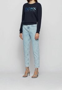 Person i mörkblå långärmad Hugo Boss-tröja, ljusblå högmidjade jeans och blå högklackade sandaler med remmar, stående.