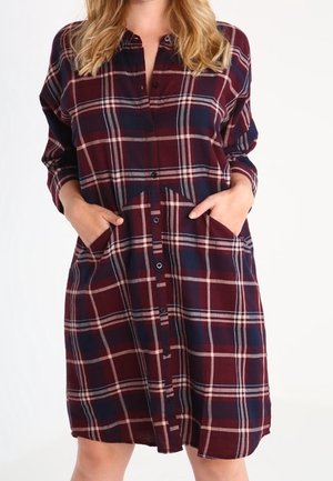 Femme portant une robe chemise à manches longues, longueur genou, avec un motif à carreaux bordeaux, marine et blanc, les mains dans les poches avant.