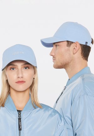 Eine Frau und ein Mann tragen passende hellblaue Harlem Soul Caps und satinierte Bomberjacken, die Frau blickt nach vorne, der Mann zur Seite.