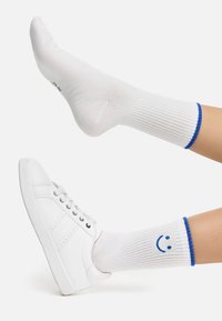 Hvide bomuldsstrømper med blå ribbede manchetter og et smiley-ansigt design. Parret med hvide sneakers, der viser glatte teksturer og et enkelt design.