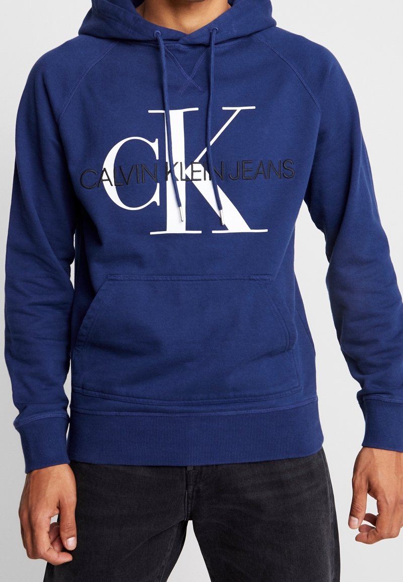 Felpa con cappuccio blu navy con un grande logo bianco "CK" e testo nero "CALVIN KLEIN JEANS." Presenta una tasca frontale e polsini a coste.