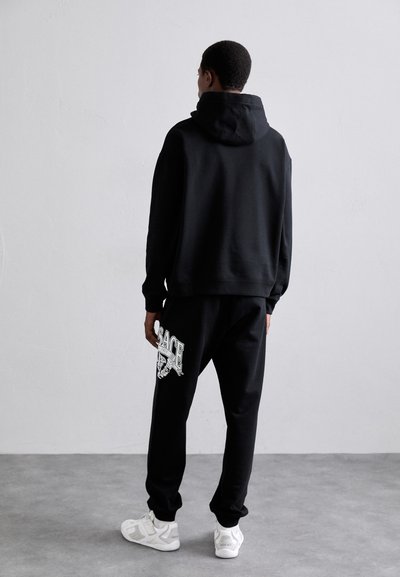 Uomo in piedi di fronte a un muro bianco, indossa un hoodie nero e pantaloni felpati con logo Versace, completando il look con scarpe da ginnastica bianche su un pavimento in cemento.