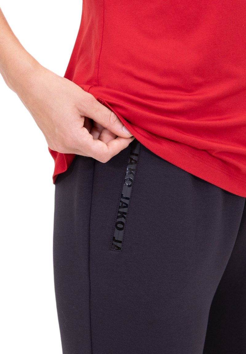 Rotes Sportoberteil mit kurzen Ärmeln, kombiniert mit dunkelblauen Hosen, die ein vertikales schwarzes Logoakzent nahe der Tasche aufweisen.