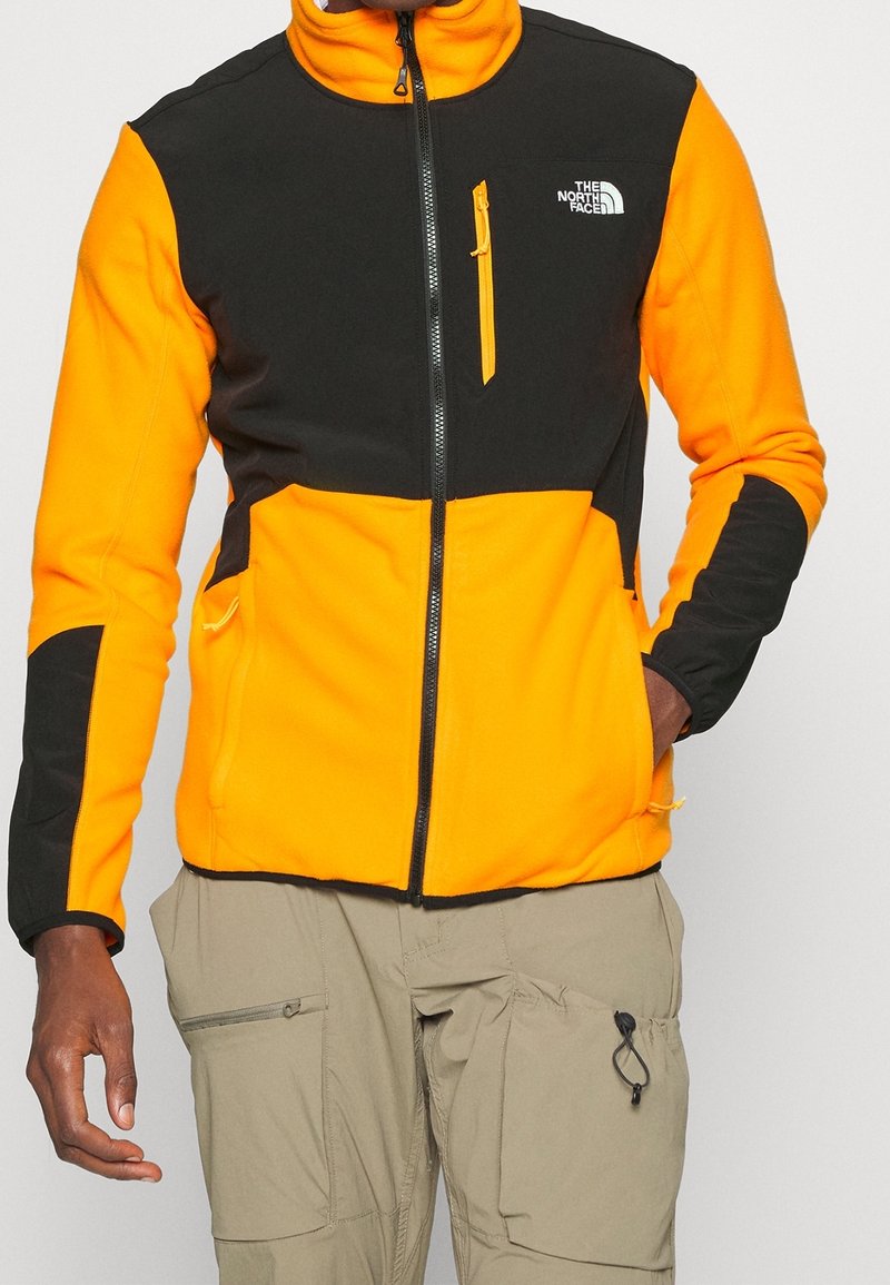 Giacca in pile con un design a blocchi di colore in arancione brillante e nero, con zip frontale, colletto alto e tasca sul petto.