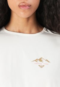 T-shirt en tissu blanc avec un graphique de montagne dorée et rose et le texte "PARADISE FOUND". Texture lisse avec un col rond.