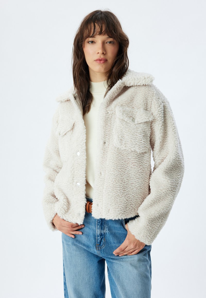 Fuzzy cremefarvet jakke med to brystlommer, krave og knaplukning. Båret over en lys sweater og parret med blå jeans.