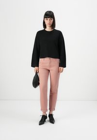 Zwarte textuurtrui, lichtroze broek, zwarte veterschoenen. Model met een kleine zwarte handtas, staand tegen een witte achtergrond.