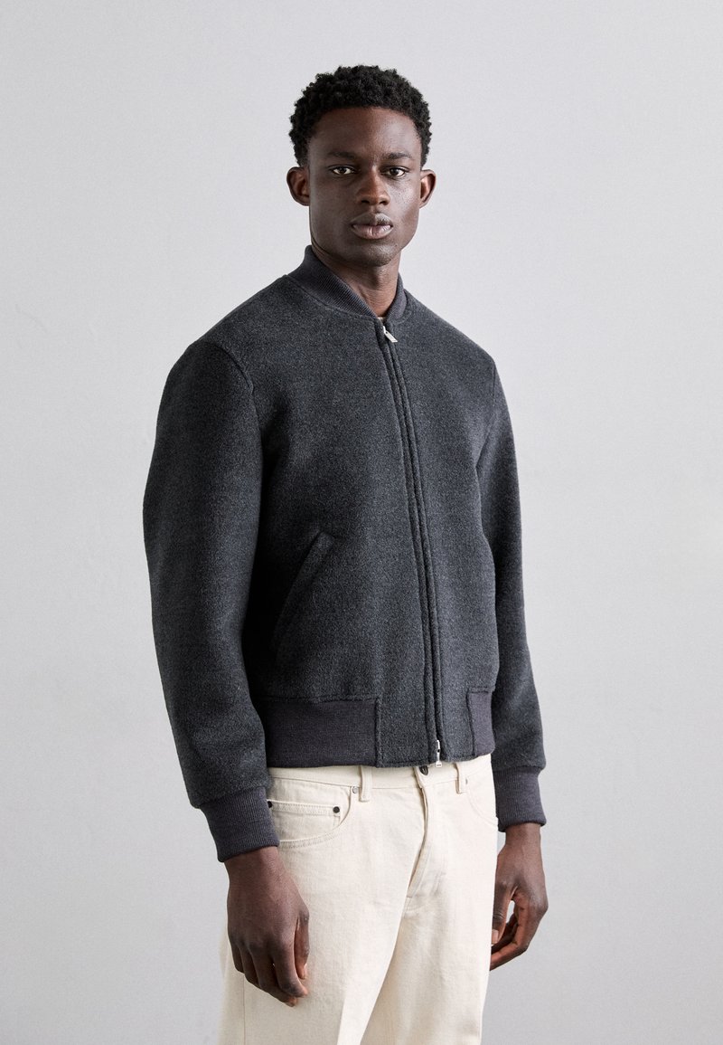 Chaqueta bomber de lana gris con cierre de cremallera en la parte delantera, puños y cinturilla de punto, y bolsillos laterales. Textura suave, diseño minimalista.