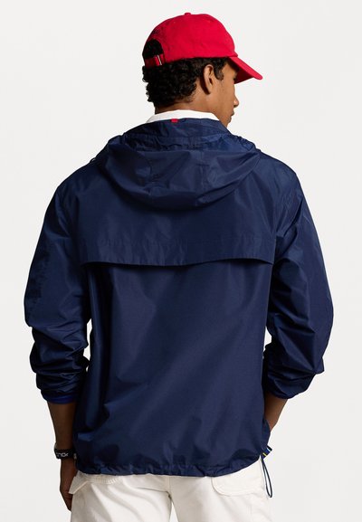 Veste coupe-vent marine avec capuche, poignets élastiques et ourlet resserré. Présente un design à dos superposé et une texture lisse et légère.