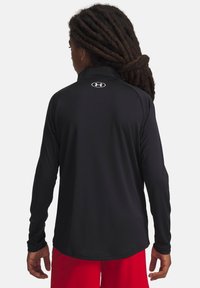 Person med lange dreadlocks iført en sort Under Armour langærmet skjorte og røde shorts, set fra bag på hvid baggrund.