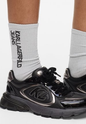 Scarpe da ginnastica nere con accenti lucidi, suole spesse e lacci neri. Calze rigate grigie con la scritta 'KARL LAGERFELD JEANS' stampata in nero.