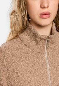 Beige Kapuzenpullover mit hochgeschlossenem Kragen, strukturiertem Stoff mit goldenen Sprenkeln und einem durchgehenden Reißverschluss auf der Vorderseite.