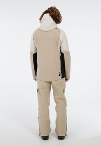 Beige waterdichte jas met zwarte accenten op de mouwen; bijpassende beige broek met zijritsen. Zachte textuur, eenvoudige ontwerpen.
