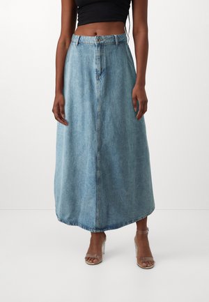 Femme portant une jupe midi en denim bleu clair taille haute avec bouton et fermeture éclair devant, associée à des sandales à talons avec brides transparentes.