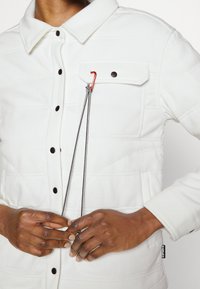 Casaco acolchoado branco com gola, apresentando um bolso no peito, botões de pressão pretos e um detalhe de cordão preto e vermelho.