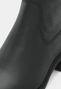 Prim-plan al unei cizme de gleznă din piele naturală neagră, cu textură netedă, cusături vizibile și un panou lateral elastic curbat.