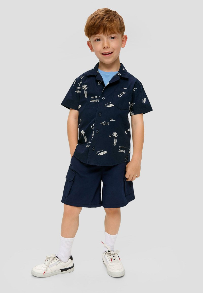 Marineblauw shirt met korte mouwen met witte prints van palmbomen en surfplanken, gecombineerd met marineblauwe cargo shorts en witte sneakers met zwarte accenten.