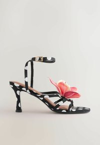 Sandale noire à talon haut avec des pois blancs, bride à la cheville avec boucle et une grande décoration florale rose sur les brides avant.