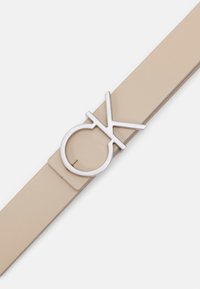 Beige leren riem met een matte afwerking en een zilveren metalen gesp met een circulair en gestileerd "CK"-logo. Soepeltextuur overal.
