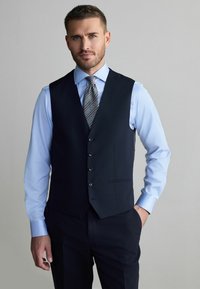 Marineblauer Weste mit fünf Knöpfen, geformtem Saum und Seitentaschen, kombiniert mit einem hellblauen Hemd und einer gemusterten Krawatte in grau, navy und weißen Streifen.