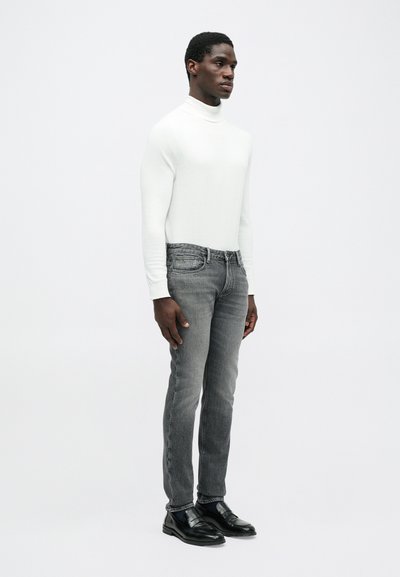 Λευκό πουλόβερ τύπου τζέρσεϊ, λεπτά γκρι τζην και μαύρα δερμάτινα παπούτσια, παρουσίαση σε standing μοντέλο με ουδέτερο φόντο.