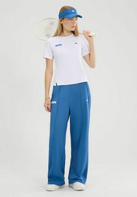 T-shirt sportivo bianco con logo blu, abbinato a pantaloni della tuta blu di taglio largo. Accessori includono un visiera blu e una racchetta da tennis.