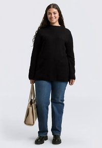 Pull en maille noire, jean bleu et bottes noires. Le mannequin tient un sac fourre-tout beige. Coupe décontractée avec manches longues et col rond.