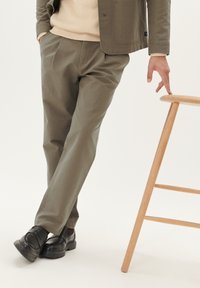Pantaloni din bumbac de culoare verde măsliniu, cu un design drept, asortați cu pantofi din piele negri, stând lângă un taburet din lemn deschis.