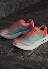 Para pomarańczowo-jasnoniebieskich butów do biegania Adidas Adizero na kamiennej nawierzchni.
