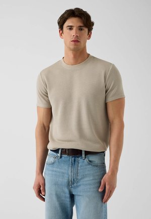 Jeune homme portant un pull en tricot beige à manches courtes rentré dans un jean bleu clair avec une ceinture noire, debout devant un fond clair uni.