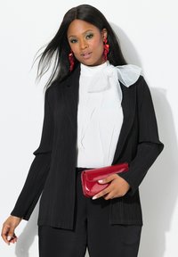Zwarte geribbelde blazer over een witte blouse met een strikdetail, gecombineerd met zwarte broek en een glanzende rode clutch. Lang zwart haar en gedurfde oorbellen.