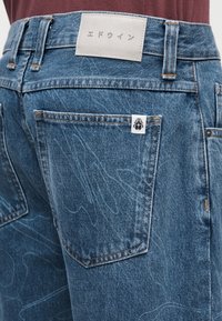 Μπλε τζιν με αφηρημένη λευκή ραφή, πίσω τσέπη με μικρή ετικέτα λογότυπου σκαθαριού και ετικέτα μέση με ιαπωνικό κείμενο.