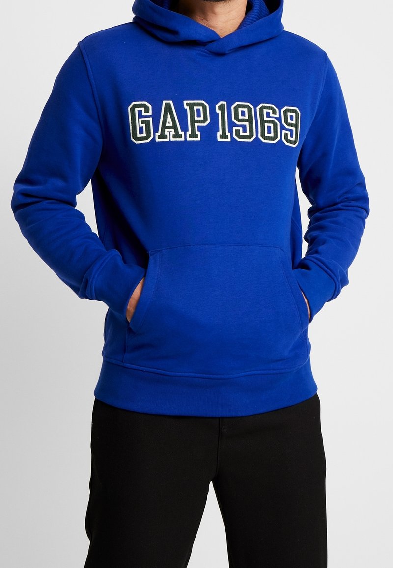 Hombre con una sudadera azul de GAP 1969 con las manos en el bolsillo frontal, combinada con pantalones negros, mostrado desde la parte media del torso hasta el cuello.