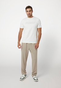 Balta trumpa rankovėmis marškinėliai su iškilusiu „GUESS“ logotipu, derinami su šviesiai smėlinėmis sportinėmis kelnėmis ir pilkai baltomis sportinėmis bateliais ant paprasto fono.